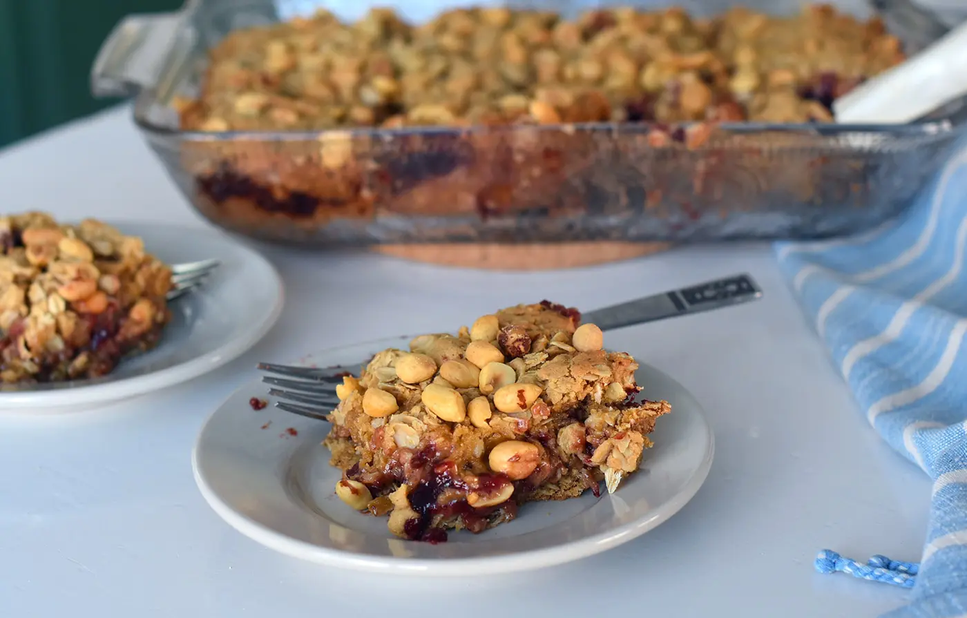 A plate with a PB & Jelly bar on it with a pan of bars in the background.