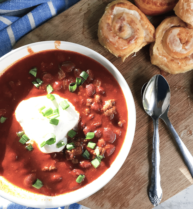 bowl of venison chili with sour cream and green onions, two spoons, and cinnamon rolls. Ground venison, beans, and tomato look hearty and warm!