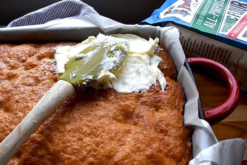 a spatula spreading cream cheese frosting on a pan of pineapple cake with a bag of Bob's Red Mill flour next to it.