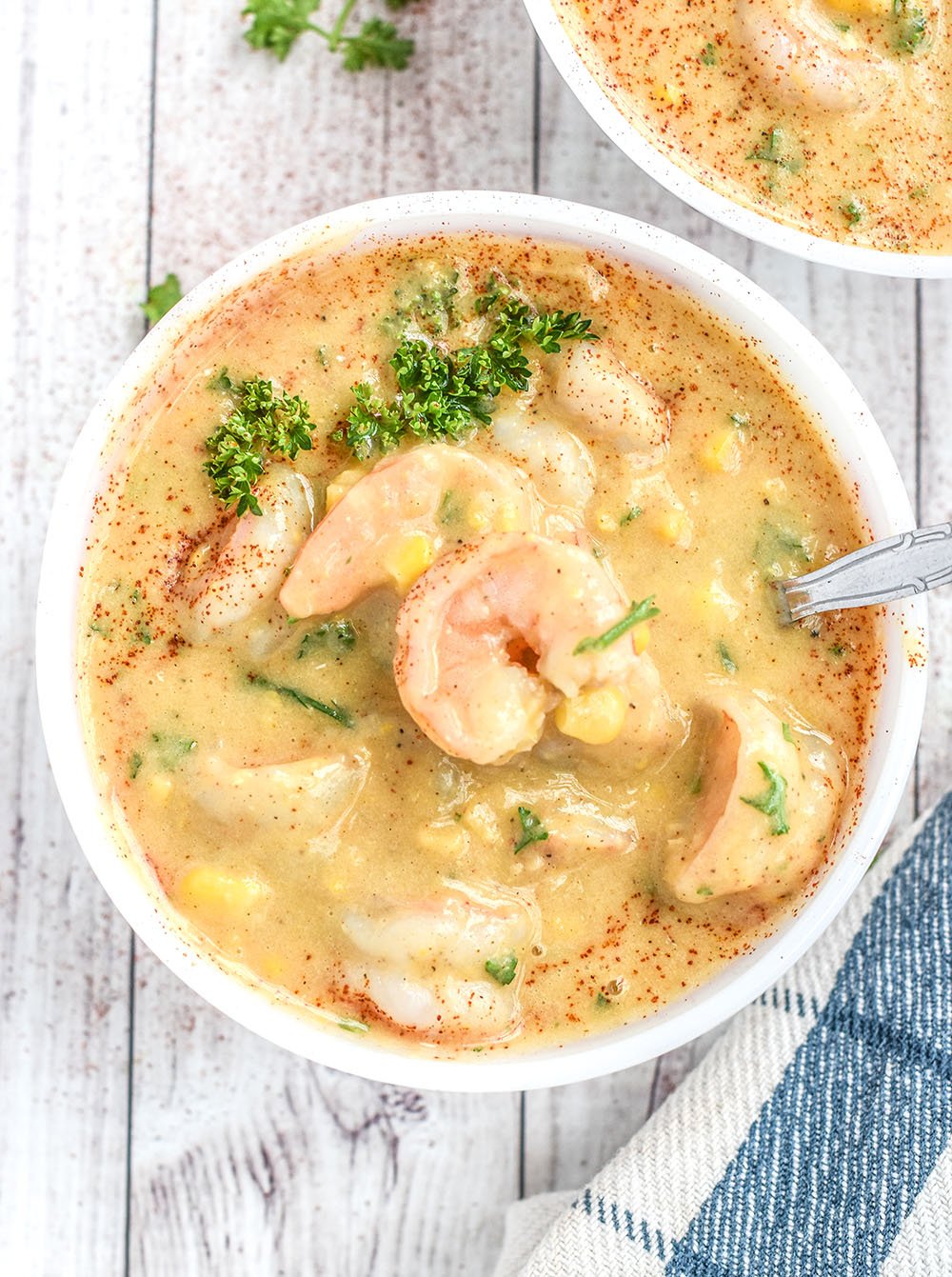 Shrimp, corn, fresh parsley, paprika, and a creamy chowder base all in a bowl with a spoon. 